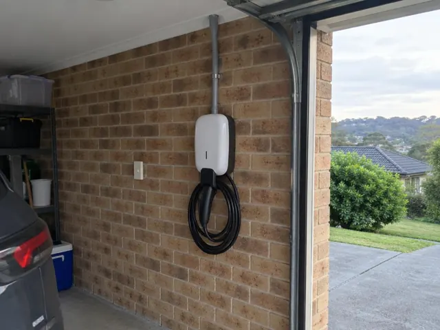 Wall-mounted EV charger with coiled cable on a brick garage wall beside a car in Kincumber