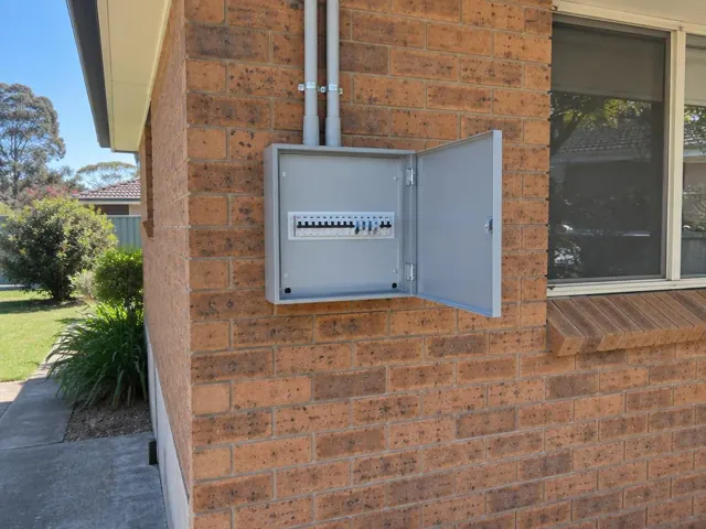 Exterior brick house wall with an open electrical switchboard cabinet beside a window in Saratoga